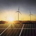 Wind turbines and solar panels sit in the foreground of a sunset.
